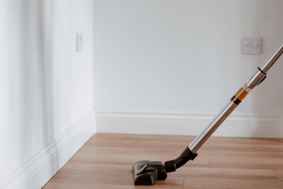 A cordless vacuum cleaner with a black base and a silver-and-transparent handle is positioned on a wooden floor near a white skirting board and an off-white wall, in a room likely prepared for surface cleaning. The room is well-lit, with a power outlet visible on the wall, and the floor appears clean and dust-free, suggesting a professional domestic cleaning environment as offered by Carpet Cleaning Colliers Wood.