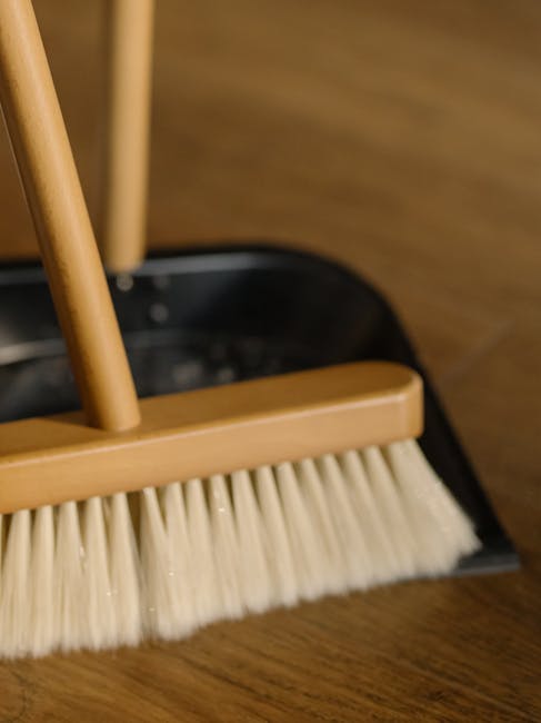 Close-up view of a broom with a light wooden handle and cream-colored bristles, positioned on a dark wooden floor next to a black dustpan. The broom and dustpan are part of a domestic cleaning setup, showcasing the tools used for surface cleaning and maintenance. The floor appears clean and well-maintained, reflecting the professional cleaning services offered by Carpet Cleaning Colliers Wood. The background features a smooth wooden surface, possibly a cabinet or wall panel, with soft lighting highlighting the cleanliness and condition of the surfaces. This image illustrates the importance of regular cleaning tools and practices in maintaining hygiene and cleanliness in residential spaces.