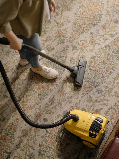 A person is vacuuming an ornate, patterned area rug with a yellow vacuum cleaner in a well-lit room, likely part of a domestic setting. The individual is wearing beige shoes and blue jeans, holding the vacuum's handle with both hands to clean the richly decorated, multicolored rug featuring floral motifs. The vacuum's black hose extends from the canister to the cleaning head, which is pressed into the carpet surface, removing dust and debris. The room's warm lighting highlights the cleanliness and condition of the rug, with the vacuum cleaner positioned on a wooden floor adjacent to the rug. This scene demonstrates a surface cleaning process, emphasizing hygiene and maintenance, and reflects the professional cleaning service provided by Carpet Cleaning Colliers Wood for home environments.