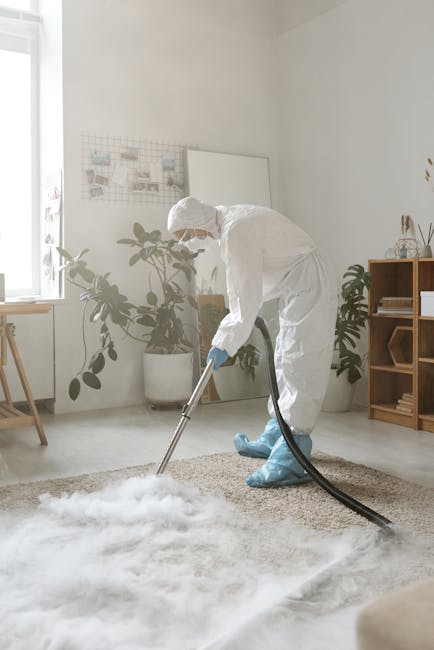 A person is vacuuming an ornate, patterned area rug with a yellow vacuum cleaner in a well-lit room, likely part of a domestic setting. The individual is wearing beige shoes and blue jeans, holding the vacuum's handle with both hands to clean the richly decorated, multicolored rug featuring floral motifs. The vacuum's black hose extends from the canister to the cleaning head, which is pressed into the carpet surface, removing dust and debris. The room's warm lighting highlights the cleanliness and condition of the rug, with the vacuum cleaner positioned on a wooden floor adjacent to the rug. This scene demonstrates a surface cleaning process, emphasizing hygiene and maintenance, and reflects the professional cleaning service provided by Carpet Cleaning Colliers Wood for home environments.