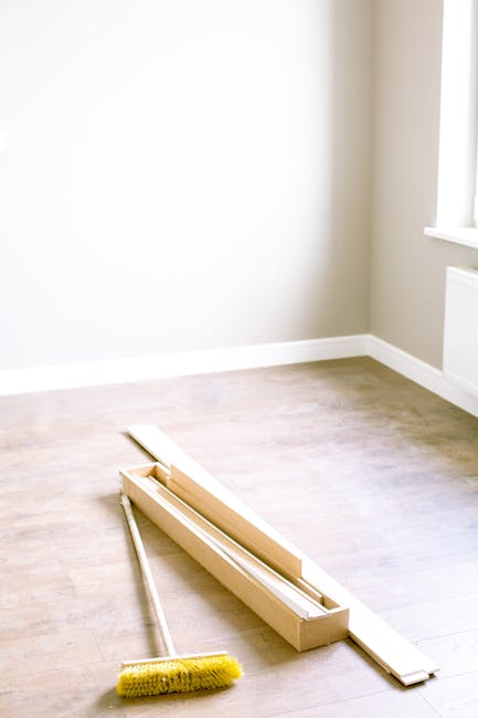 A bright, empty room with light wooden flooring and beige walls, featuring a white baseboard along the bottom edge. Natural light streams in through a nearby window, illuminating the space. On the floor, there are cleaning tools including a long, narrow wooden skirting board or trim, a yellow scrub brush, and a small rectangular box possibly containing additional cleaning accessories. The room appears freshly cleaned and ready for finishing touches, exemplifying surface cleaning and preparation for deep or domestic cleaning services provided by Carpet Cleaning Colliers Wood. The space is tidy, with no visible dust or dirt, highlighting a focus on hygiene and proper maintenance of the flooring and surfaces.