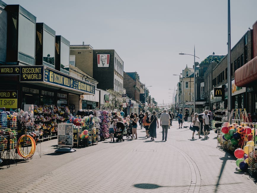 A bustling pedestrian street on Colliers Wood High Street lined with various small retail stores and stalls, including a floral and decorative items vendor on the left and balloon vendor on the right. The street is paved with light-colored bricks, and the storefronts feature modern and traditional architecture with large windows and signage. Shoppers, some pushing strollers and carrying shopping bags, walk along the wide, clean pavement under bright daytime skies. Visible shop signs include 'Discount World' and 'KFC'. Streetlights and a mix of older and newer buildings enhance the vibrant commercial atmosphere, with ample natural light making the scene appear bright with minimal shadows. As a professional cleaning specialist, Carpet Cleaning Colliers Wood ensures the cleanliness of such high-traffic public spaces through effective surface cleaning, deep cleaning, and regular sanitisation of the pavements and storefront exteriors to maintain hygiene standards.