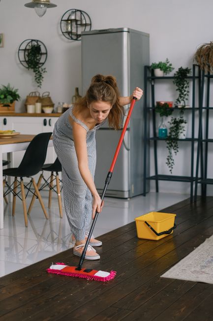 A young woman with light brown hair tied back, dressed in a striped jumpsuit, is performing surface cleaning in a modern kitchen. She is using a red-handled mop with a pink cleaning pad to scrub a dark wooden floor, which appears freshly mopped and glossy. The kitchen features a stainless steel refrigerator, black shelving units with green plants and kitchenware, and a table surrounded by black chairs with wooden legs. A yellow cleaning bucket is placed on the floor nearby. Bright, natural lighting illuminates the space, emphasizing the cleanliness and hygiene of the area. This scene is representative of domestic cleaning services provided by Carpet Cleaning Colliers Wood, focusing on deep cleaning and surface sanitisation of residential surfaces.