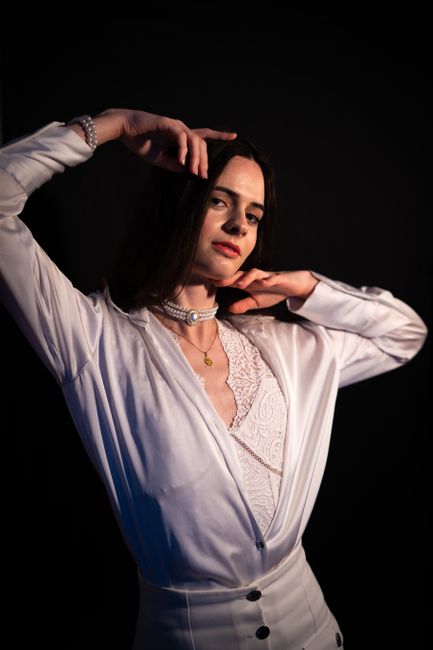 A young woman with shoulder-length dark hair and fair skin, dressed in a white satin blazer and lace top, stands against a dark background. She is adjusting her hair with both hands, one hand near her